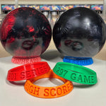 Two large bowling balls and four colorful wristbands with text on a bowling alley background.